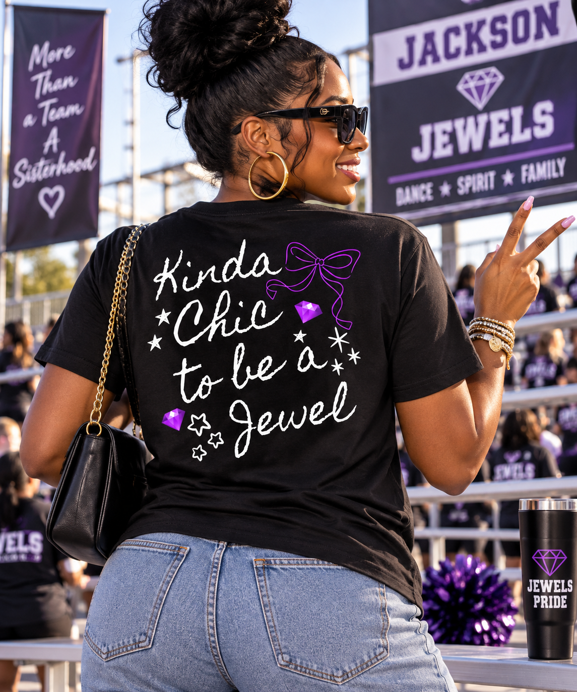 A woman photographed from behind at an outdoor event, wearing a black graphic T-shirt with the phrase 'Kinda Chic to be a Jewel' in white script and purple diamond/bow graphics. The background shows bleachers, crowd, and banners, including one that reads 'Jackson Jewels'.