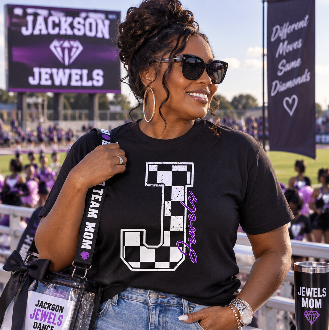 A woman at a football stadium wearing a black graphic t-shirt with a large white and purple letter 'J' design, paired with light-wash ripped jeans. She is carrying a clear shoulder bag with a purple and white 'Jackson Jewels Dance Team Mom' strap, and a matching black tumbler cup is visible in the foreground.