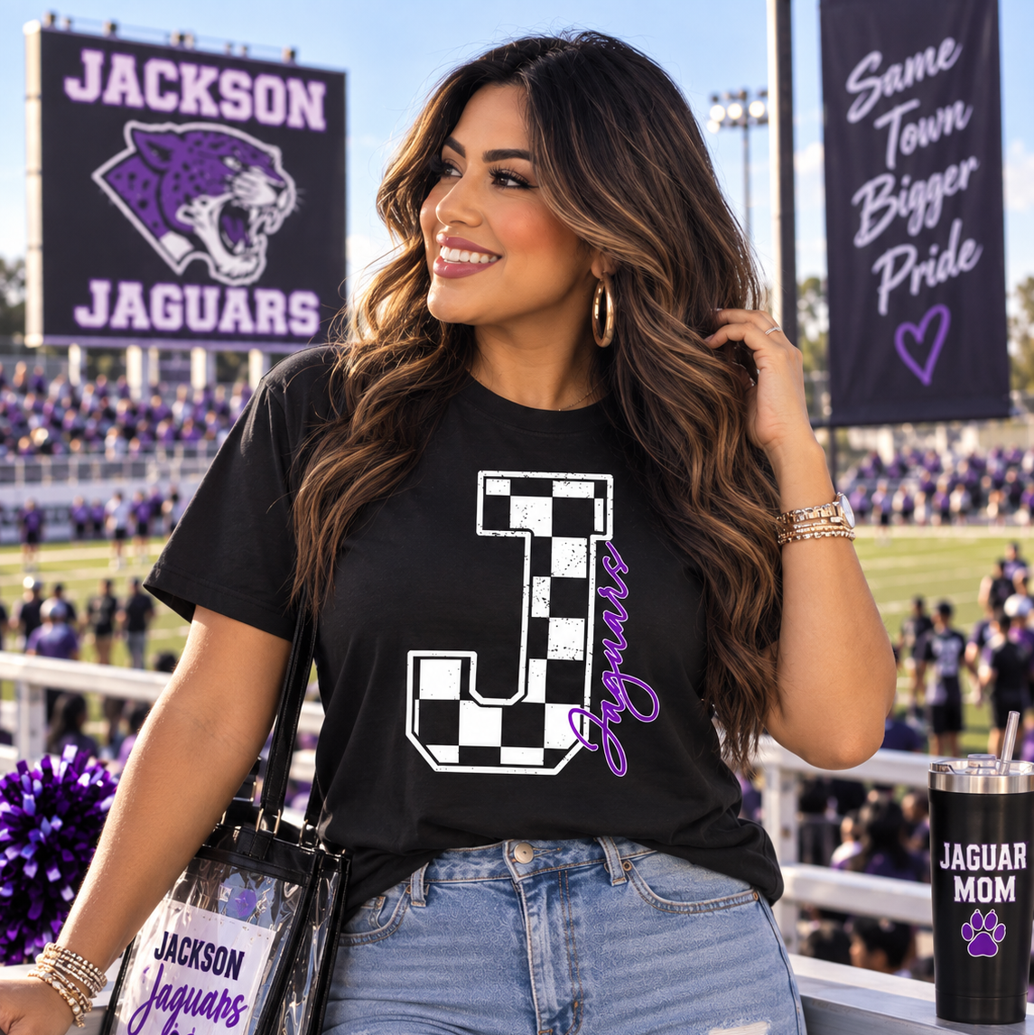 A woman stands at a sports event wearing a black graphic t-shirt with a large checkered letter 'J' and the word 'Jaguars' in purple script. She is also wearing ripped blue jeans and carrying a clear shoulder bag with 'Jackson Jaguars' text and a paw print.