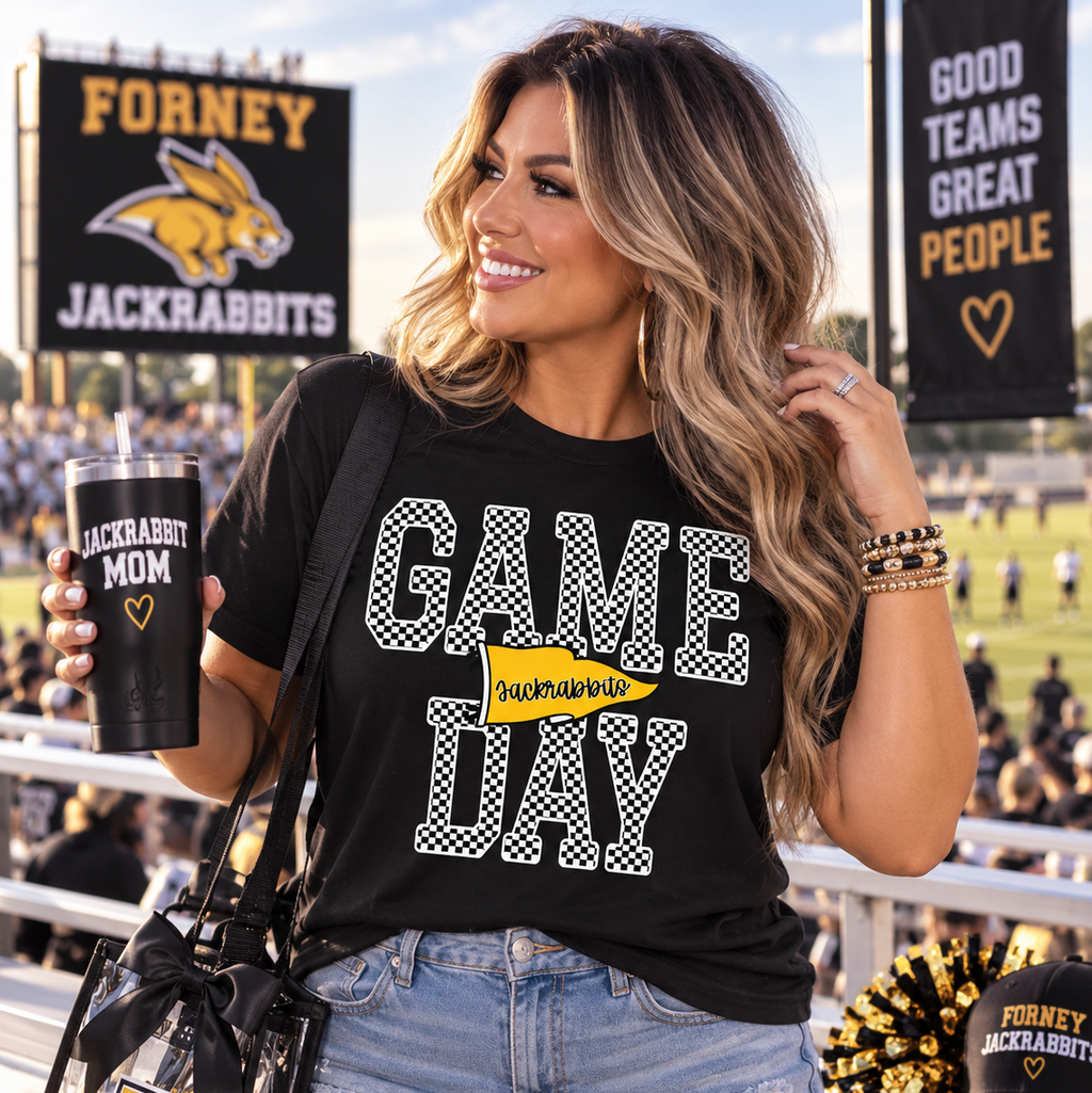 A woman stands in a stadium holding a black tumbler and wearing a black graphic tee that reads 'GAME DAY JACKRABITS' with a yellow pennant accent. She has a clear shoulder bag with a gold 'F' and a black cap on the bench beside her.