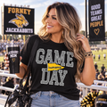 A woman stands in a stadium holding a black tumbler and wearing a black graphic tee that reads 'GAME DAY JACKRABITS' with a yellow pennant accent. She has a clear shoulder bag with a gold 'F' and a black cap on the bench beside her.