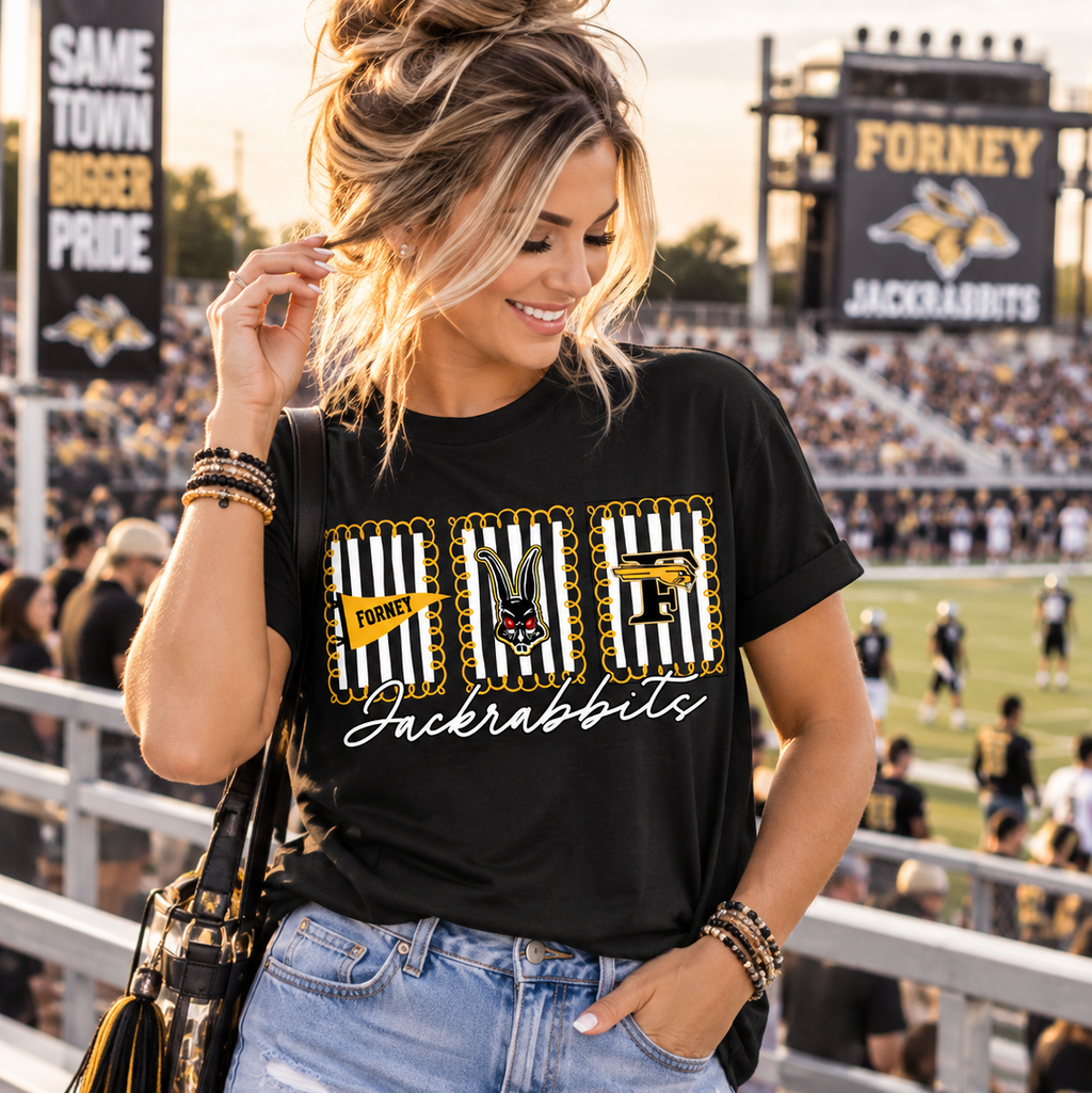 A woman stands at a football stadium wearing a black graphic t-shirt with 'NUG Jackrabbits' in a striped and yellow design, paired with light-wash denim shorts and a black tassel shoulder bag.