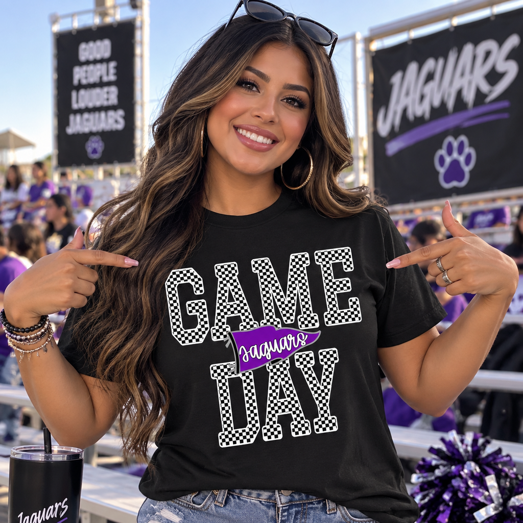 A woman wearing a black graphic t-shirt with 'GAME DAY JAGUARS' printed on the front, sitting at a bleachers in a stadium. She is pointing at the shirt with both hands, wearing denim shorts and a black tumbler cup.