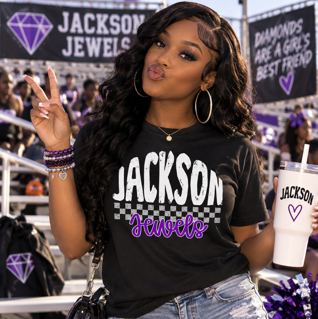 A woman posing in a black graphic t-shirt that reads 'Jackson Jewels' in white and purple, paired with distressed denim shorts. She is holding a matching white tumbler cup with 'Jackson Jewels' branding, set against a stadium background with fans and banners.