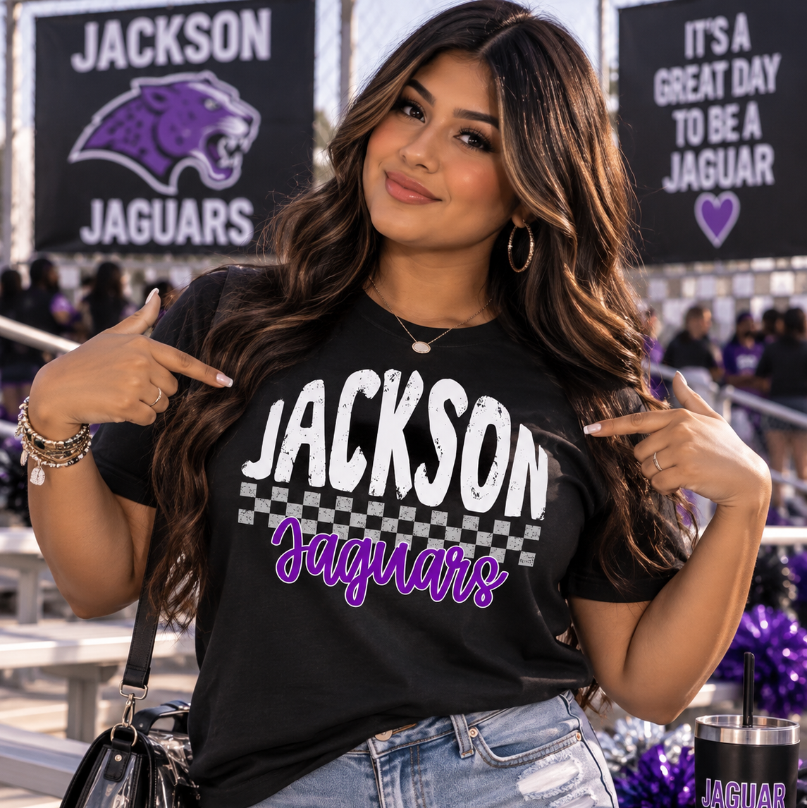 A woman wearing a black graphic t-shirt with 'Jackson Jaguars' printed in white and purple, paired with distressed denim shorts, posing in a stadium setting with team banners in the background.
