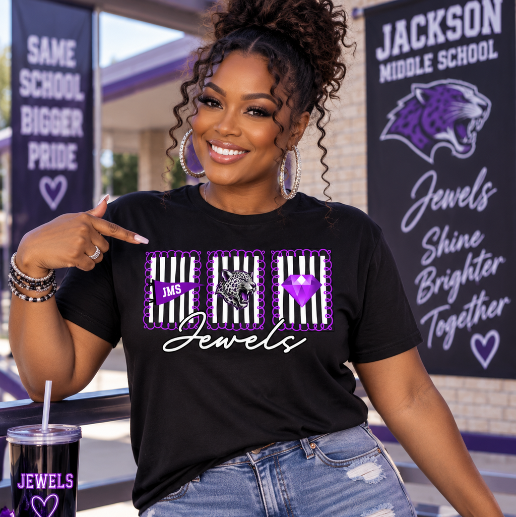 A woman wearing a black graphic t-shirt with purple and white designs, posing in front of Jackson Middle School banners. The shirt features three panels with black and white stripes, purple hearts, and the word 'Jewels' in white script.