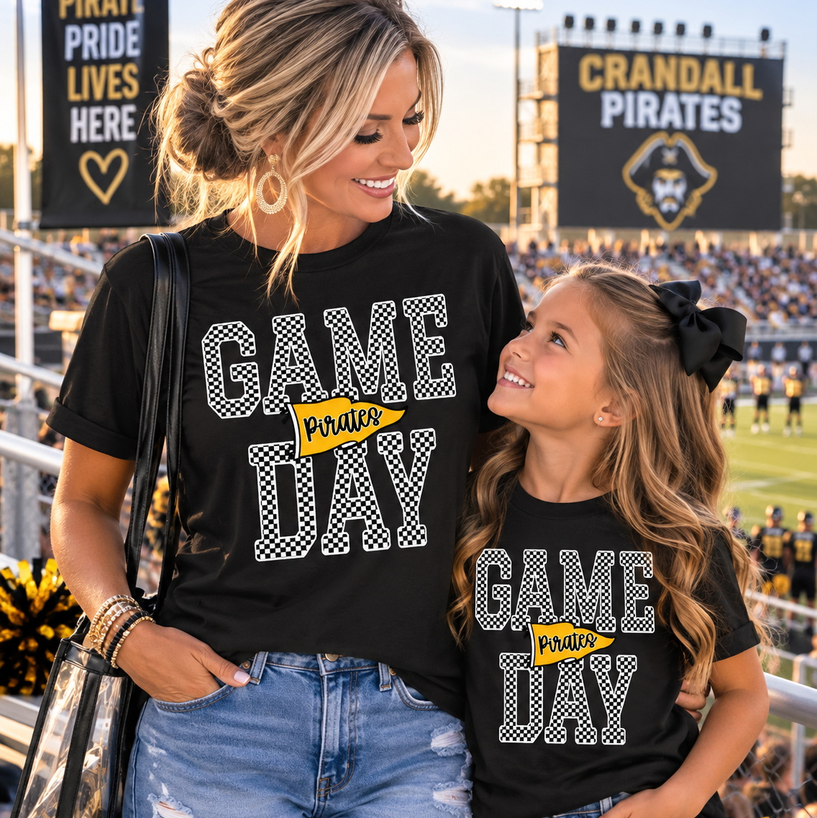 A woman and a young girl wearing matching black Crandall Pirates 'Game Day' graphic t-shirts, standing in a stadium with cheerleaders and stadium lights in the background.