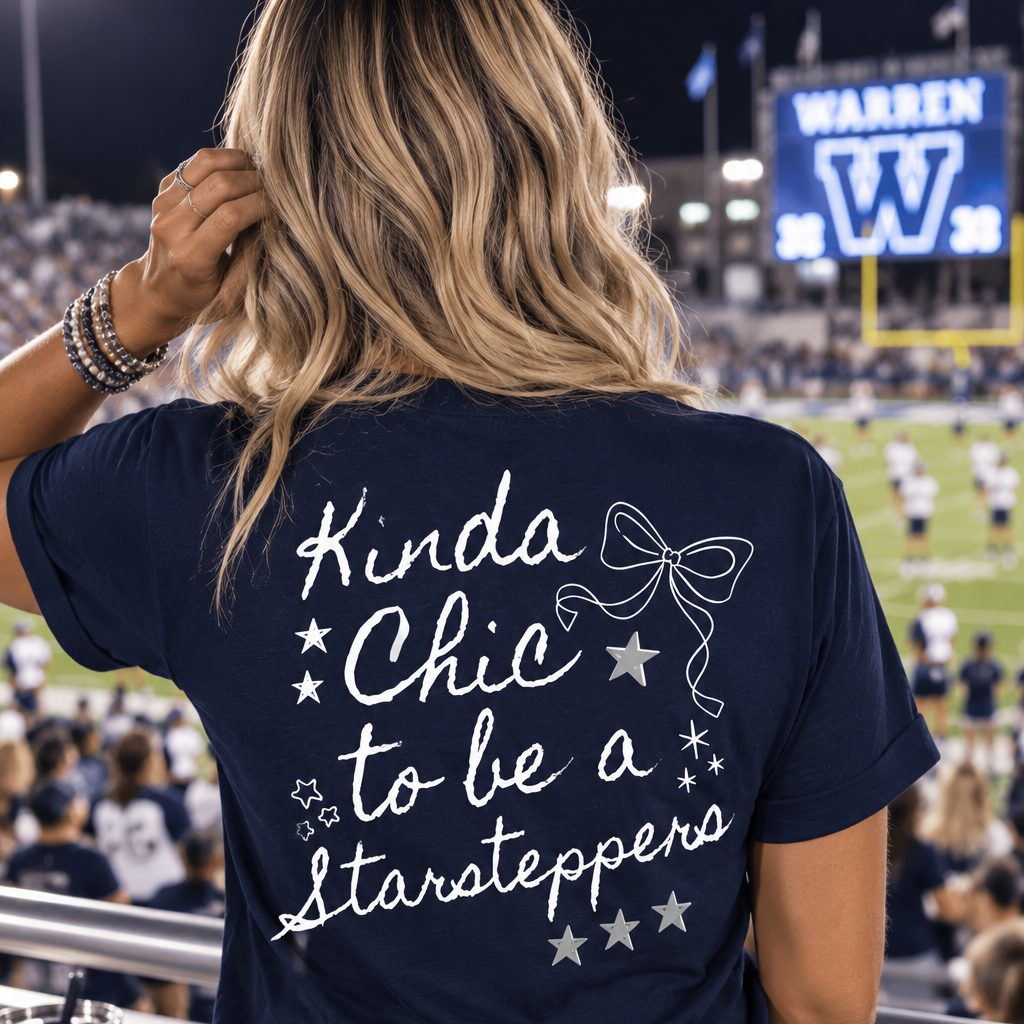A woman viewed from behind wearing a navy blue t-shirt with white text that reads 'Kinda Chic to be a Starsteppers' and a bow graphic, standing at a stadium night game.