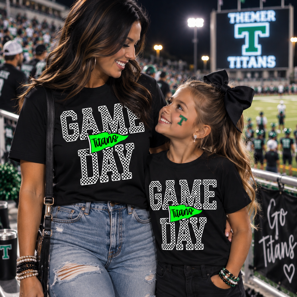A woman and a young girl wearing matching black graphic T-shirts that read 'GAME DAY Titans' in white and green, standing together at a football stadium at night. The girl has a green 'T' face paint on her forehead and wears a black bow in her hair.