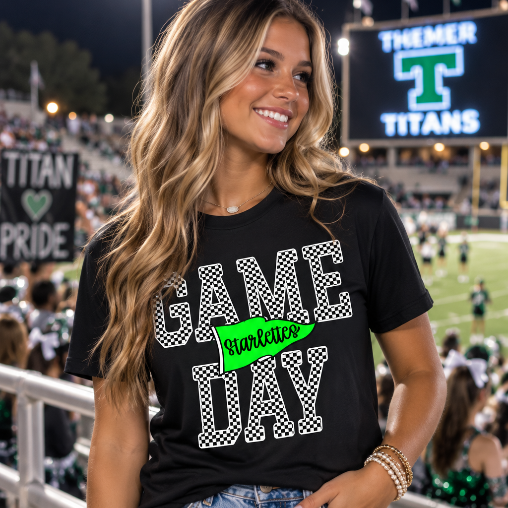 A woman stands in a stadium wearing a black graphic T-shirt that reads 'GAME DAY' in white patterned letters with a bright green 'turkeyday' banner across the front, paired with light blue denim shorts.