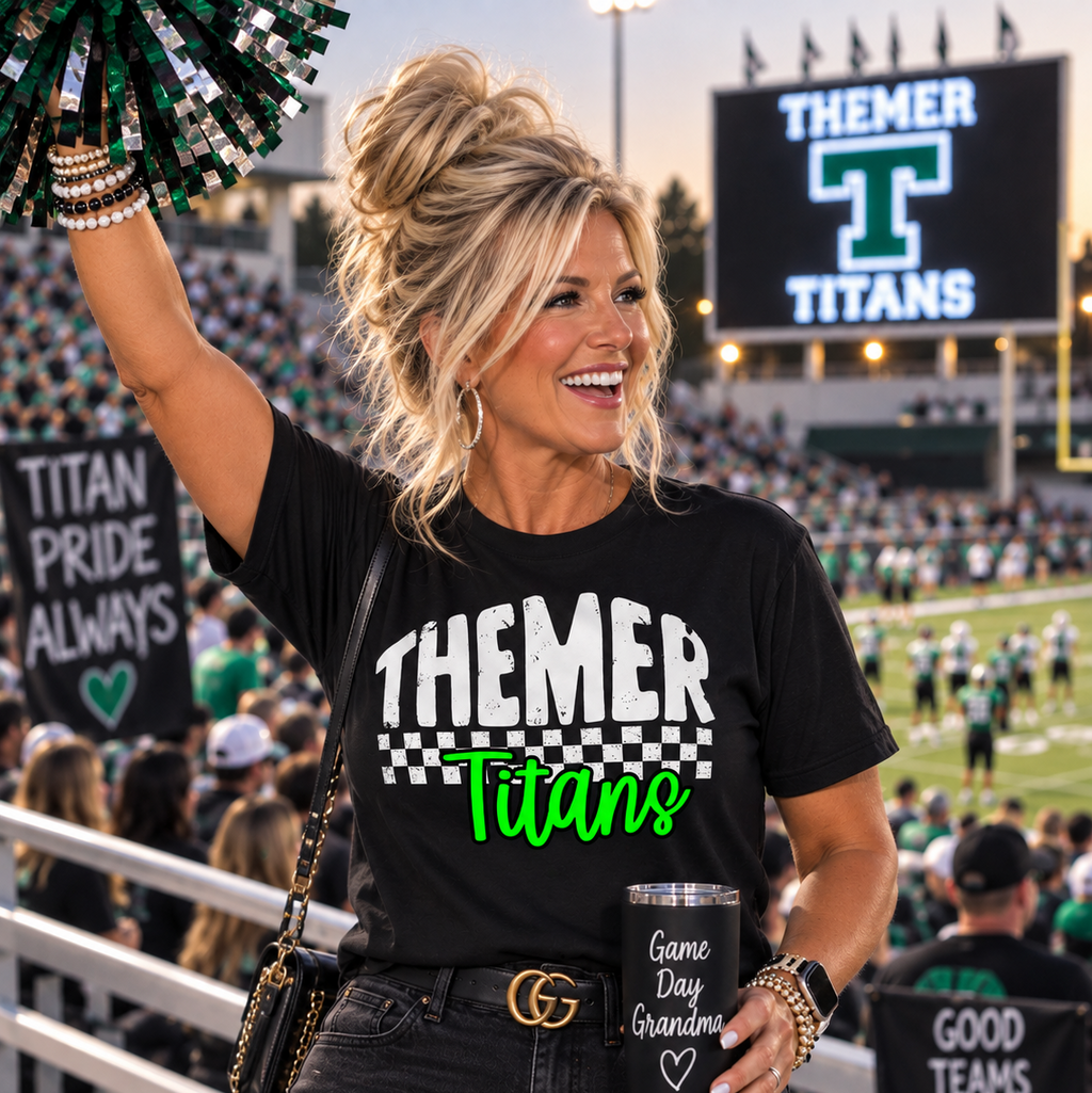A woman cheerleader at a football stadium wearing a black graphic t-shirt that reads 'THEMER Titans' in white and green, paired with a black tumbler and jeans.