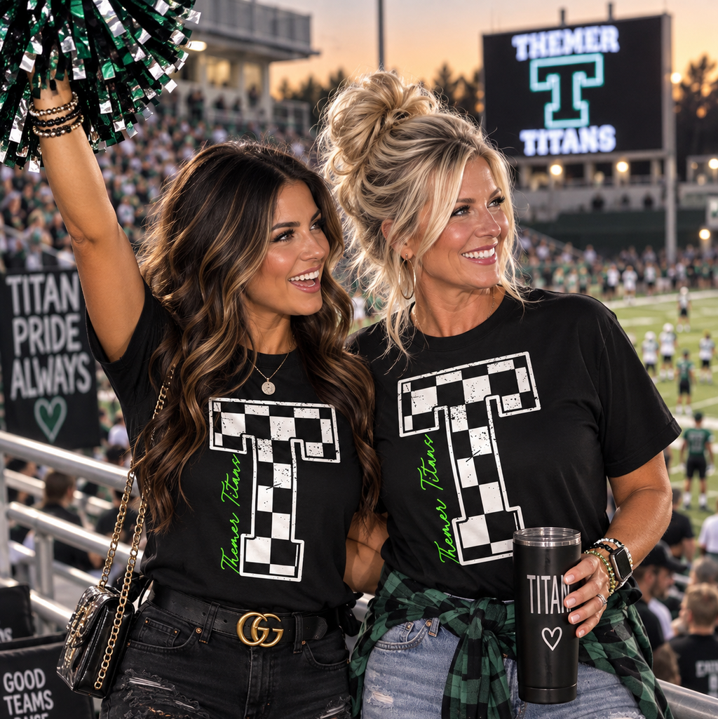 Two women standing in a stadium crowd wearing matching black T-shirts with a large white checkerboard 'T' graphic and green 'Team Titans' text. One holds a green-and-black pom-pom, and the background shows a football field and large team scoreboard.
