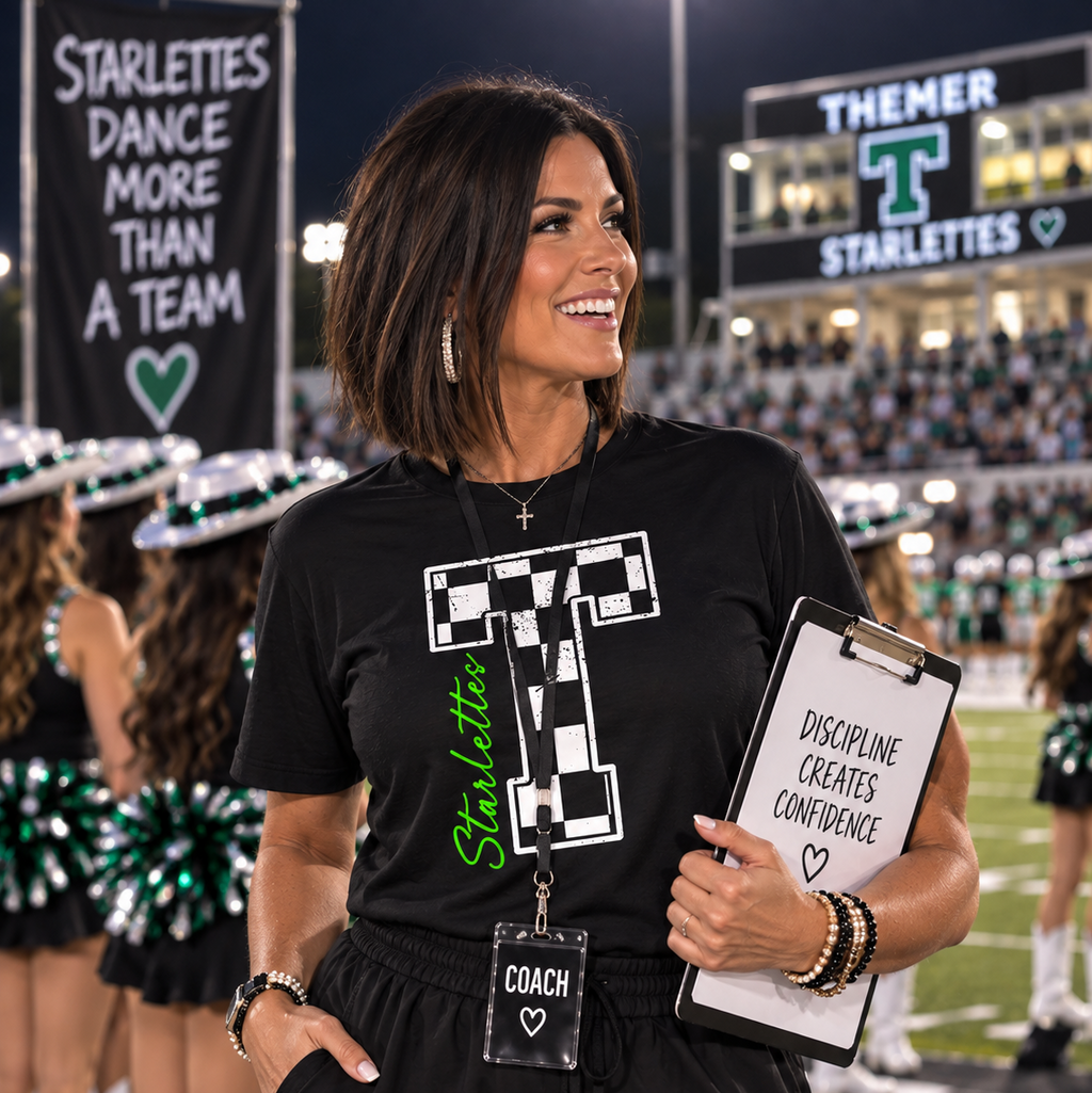 A woman on a football field at night wearing a black graphic T-shirt with a large white checkerboard letter T and green 'Starlettes' text, holding a clipboard. Cheerleaders and stadium signage are visible in the background.
