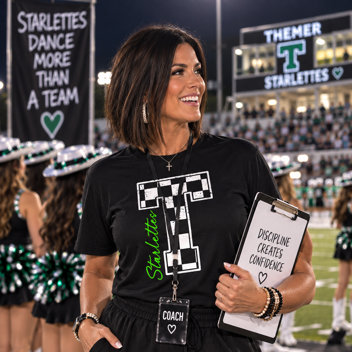 A woman on a football field at night wearing a black graphic T-shirt with a large white checkerboard letter T and green 'Starlettes' text, holding a clipboard. Cheerleaders and stadium signage are visible in the background.