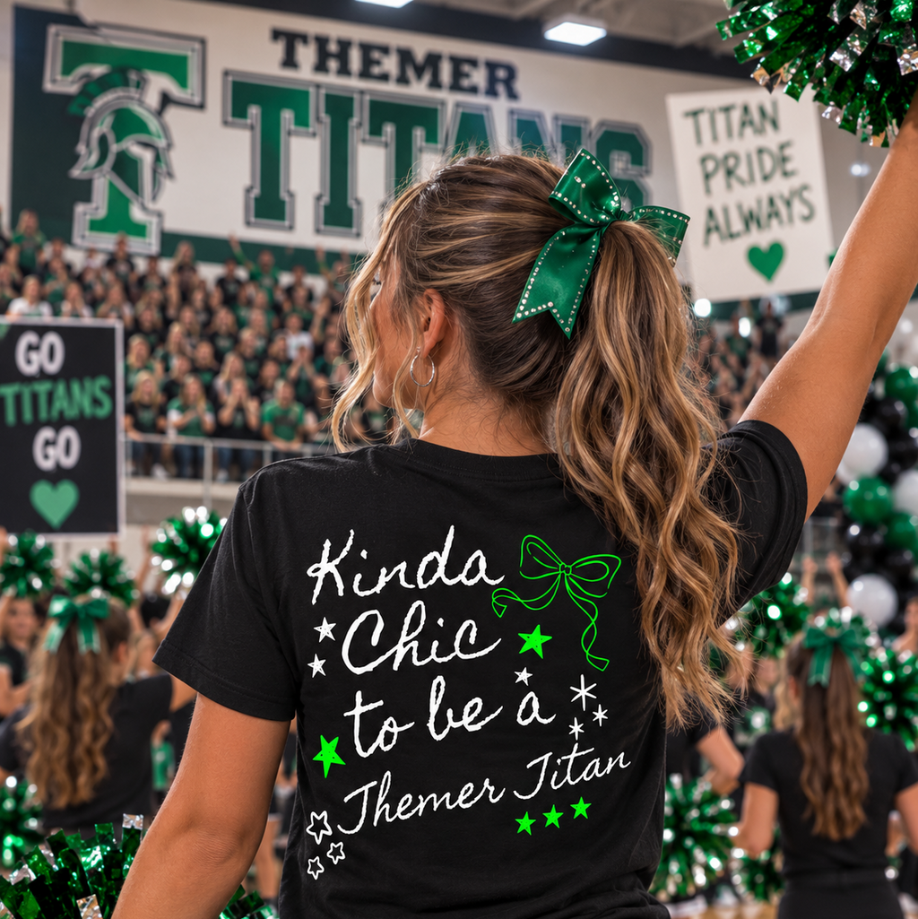 A woman seen from behind wearing a black graphic T-shirt with green and white text reading 'Kinda Chic to be a Themer Titan' and a green bow in her ponytail, standing in a cheerleader setting with Themer Titans signs in the background.