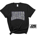 A black short-sleeve t-shirt with 'JAGUARS' printed across the chest in a textured gray and white pattern with a purple outline. The shirt is styled with a knot at the bottom hem and features a 'J29 SPIRIT' logo overlay on the bottom right.